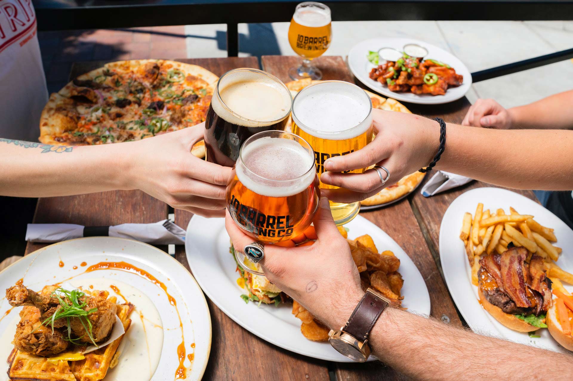 Photo of a group of people's hands putting their beer glassses together over a table full of food.